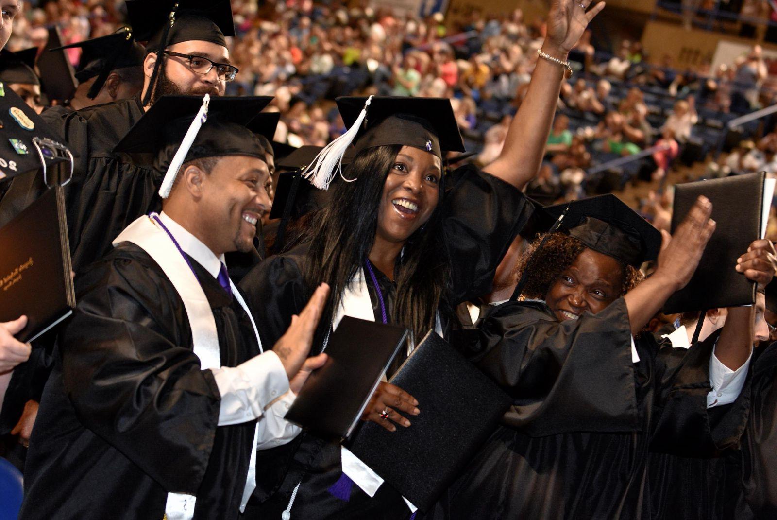 A forest green, hyperlinked, button labeled 技术信用证书 in white, bolded, underlined text with an image of a crowded gathering of graduates and family focusing on four graduates wearing black graduate caps and gowns with white tassels and stoles: an African American female smiles while holding a black diploma/degree holder up with her left hand placed on the side, to her left an African American female smiles and raises her right hand wearing a metal bracelet while holding her black degree/diploma holder in her left hand, an African American male stand smiling and holding his black diploma/degree holder in his right hand with his left hand held up behind it, and a Caucasian male with a  black beard and glasses stands behind and to the right of those three.